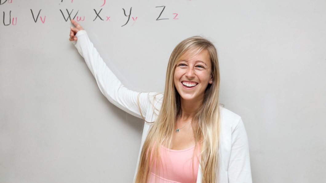 A teacher stands at a whiteboard. She is smiling.